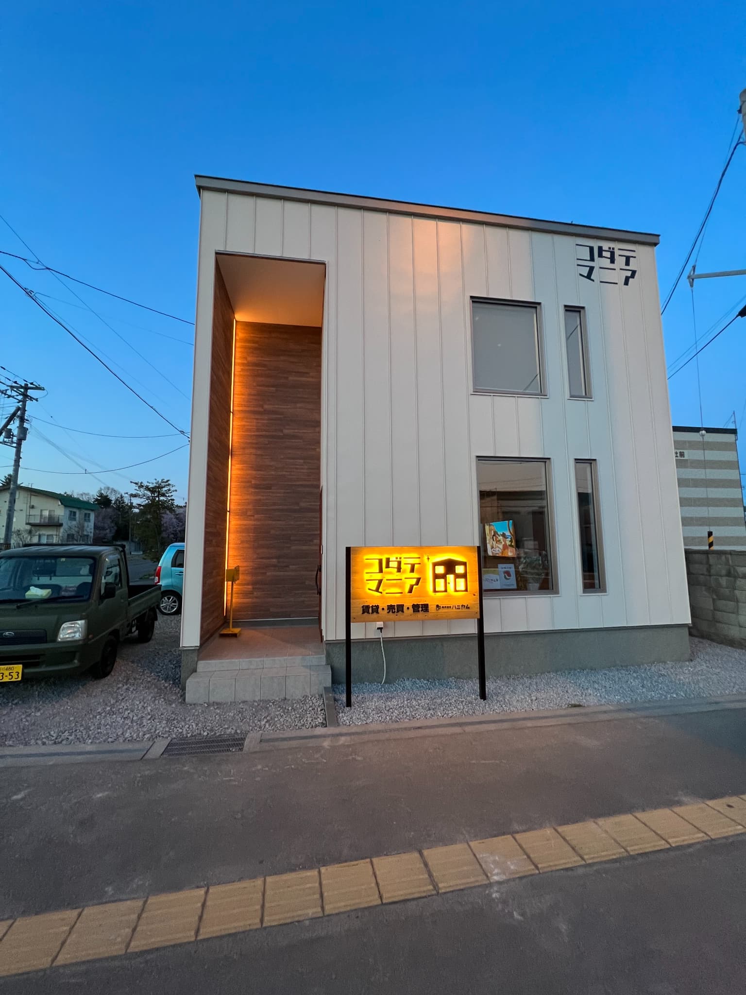 旭川市花咲町の新築店舗兼オフィスの建物全体写真。夕暮れの青空の下、白い箱型のモダンな外観デザインと、手前の駐車場スペースが写っている