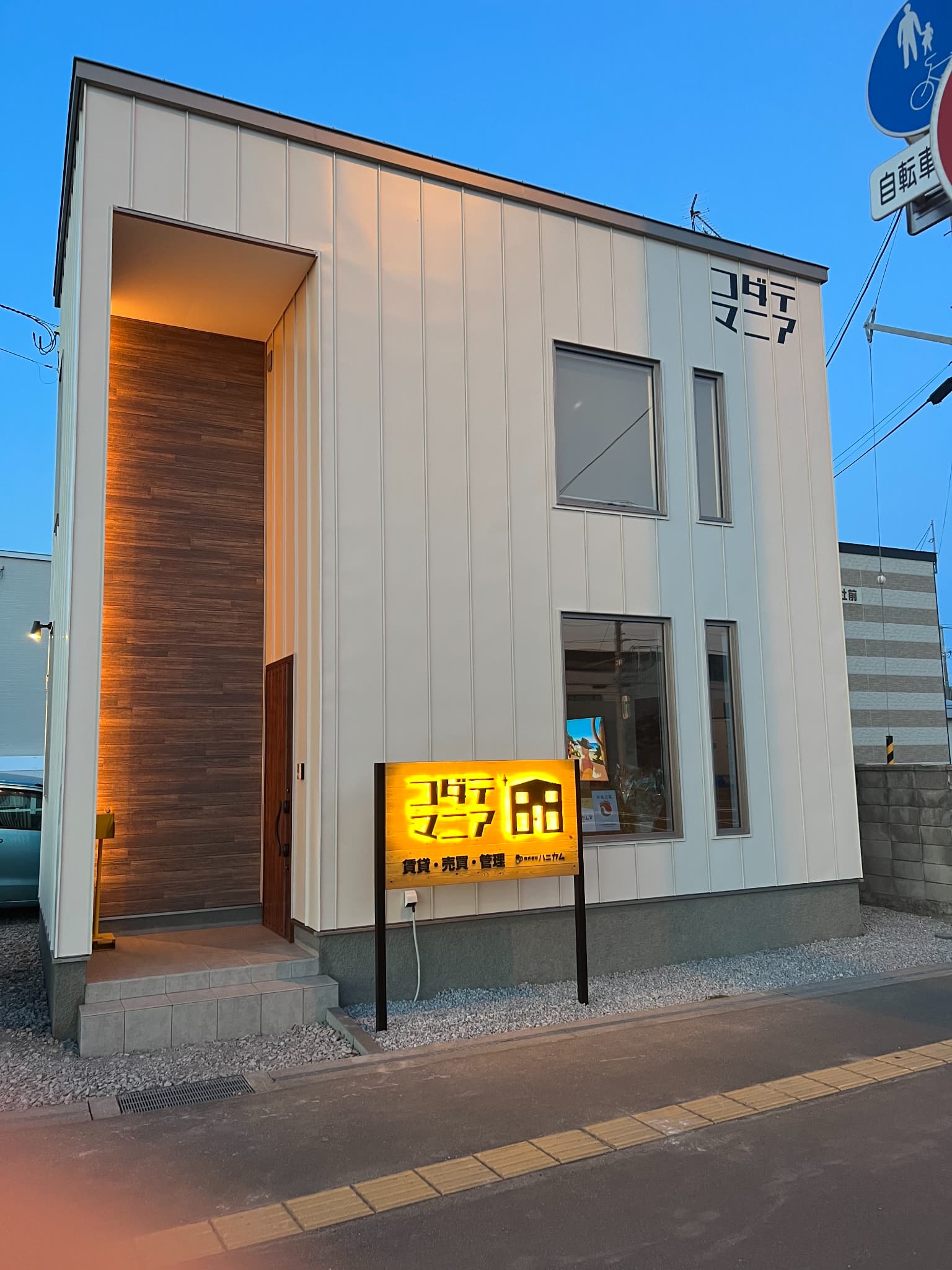北海道旭川市花咲町に新築された不動産店舗「コダテマニア」の夕暮れ時の外観。白いガルバリウム外壁と、暖色系のライトで照らされた木目調のエントランスポーチ、黄色く光る電飾看板が特徴的。