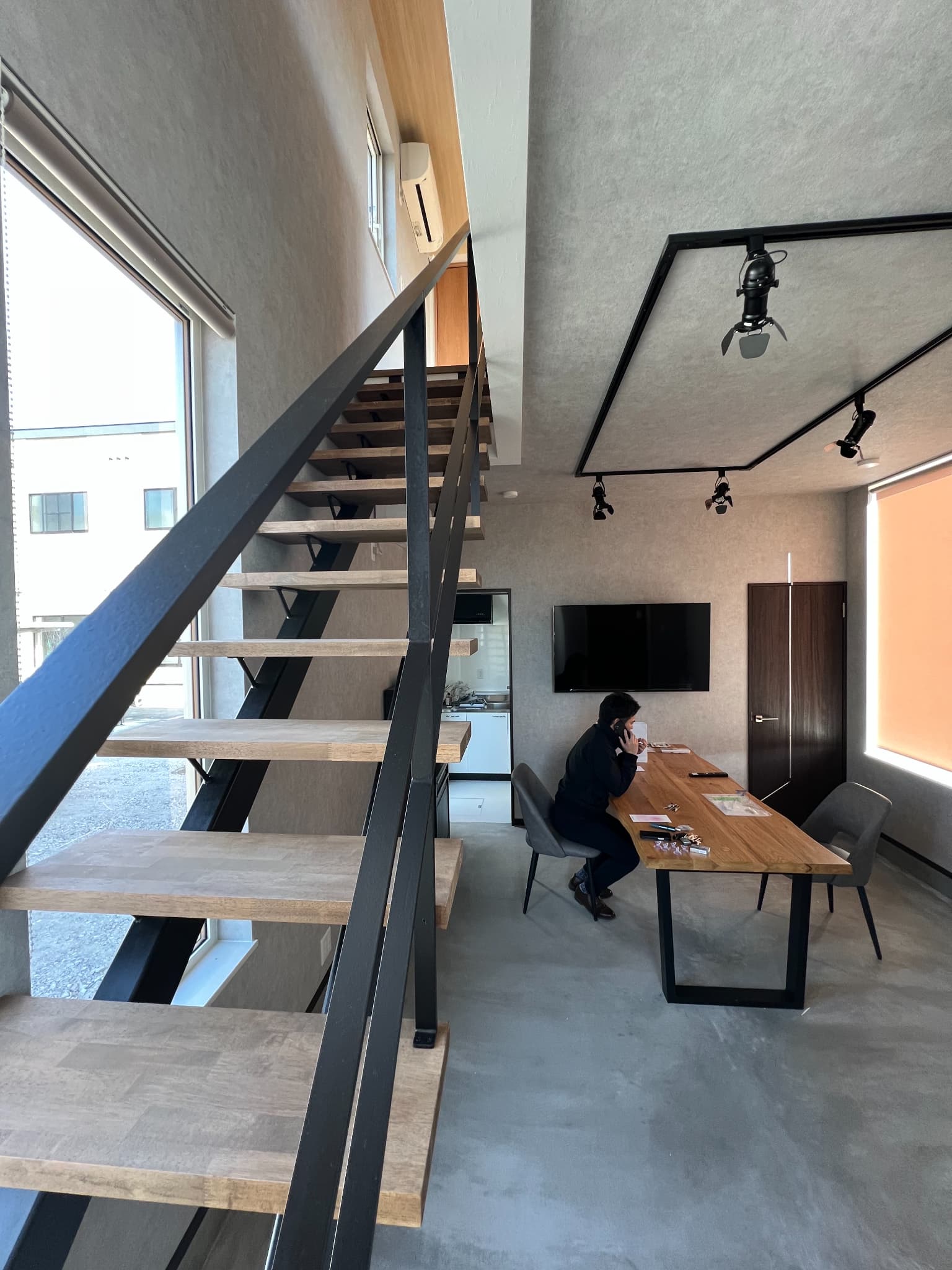 旭川市の店舗新築施工事例の内観写真。コンクリート調の壁面に映える黒い鉄骨スケルトン階段と、大きな窓がある開放的な打ち合わせスペース。インダストリアルなデザインのオフィス空間。